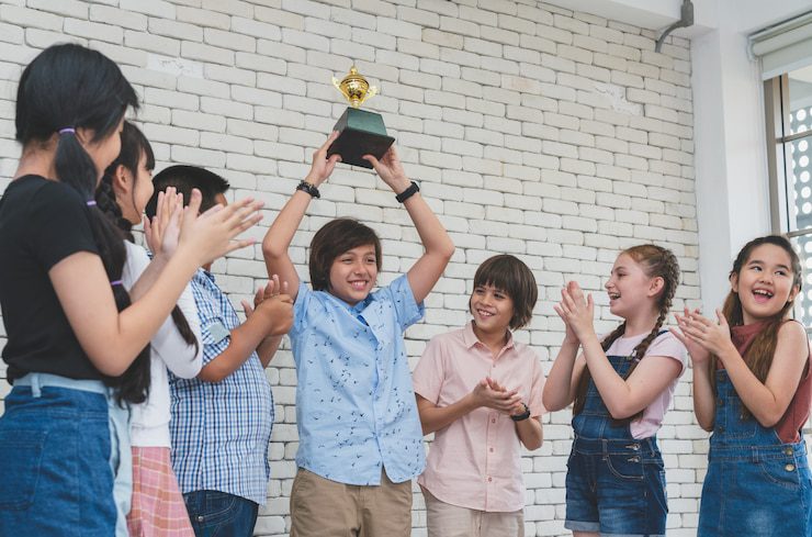 group-children-congratulated-happy-boy-holding-trophy-cup-award-his-winning-classroom_34755-304